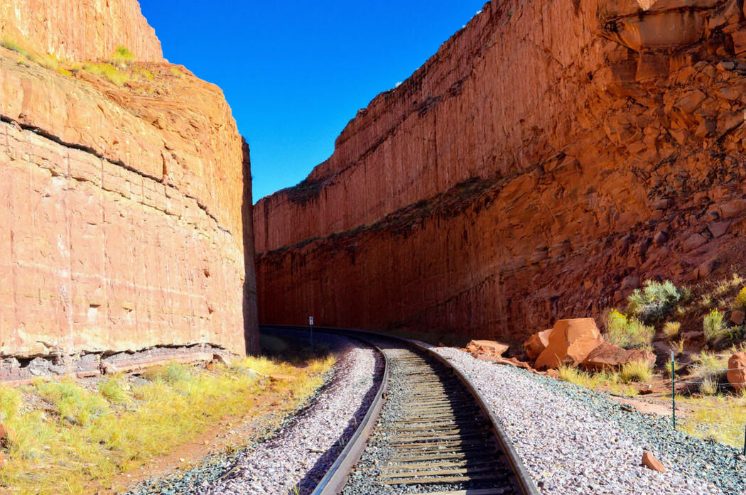 Just Around The Bend by CJ Cusinato Railroad tracks curve through a narrow canyon with towering red rock walls under a vibrant blue sky. Sparse desert vegetation and scattered boulders line the path, highlighting the contrast between rugged nature and human infrastructure.