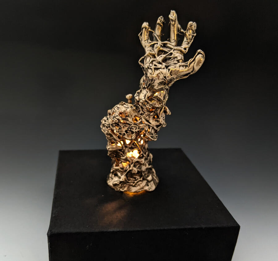 Night Light by Eva Xhan A sculptural night light featuring a metallic, abstract form of a hand emerging from a textured base resembling intertwined faces and organic shapes. Warm light glows through openings, casting intricate shadows against a dark background.