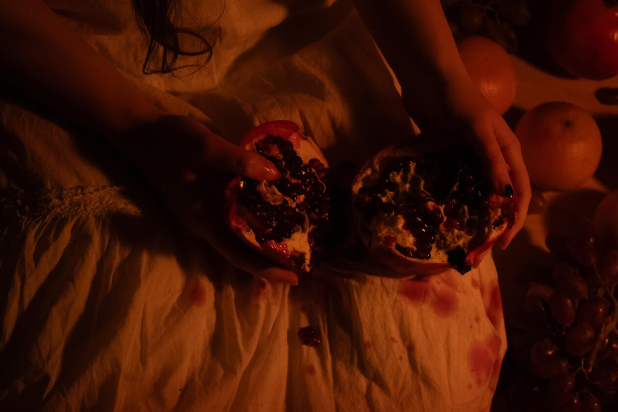 Sapphic by Ella Bertsch A dimly lit scene of hands holding a split pomegranate over a white dress stained with juice. Surrounding fruits and soft shadows create a warm, intimate atmosphere, evoking themes of sensuality, nature, and quiet reflection.